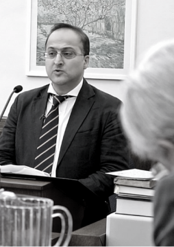 Person in a suit speaking into a microphone at a desk with papers and books