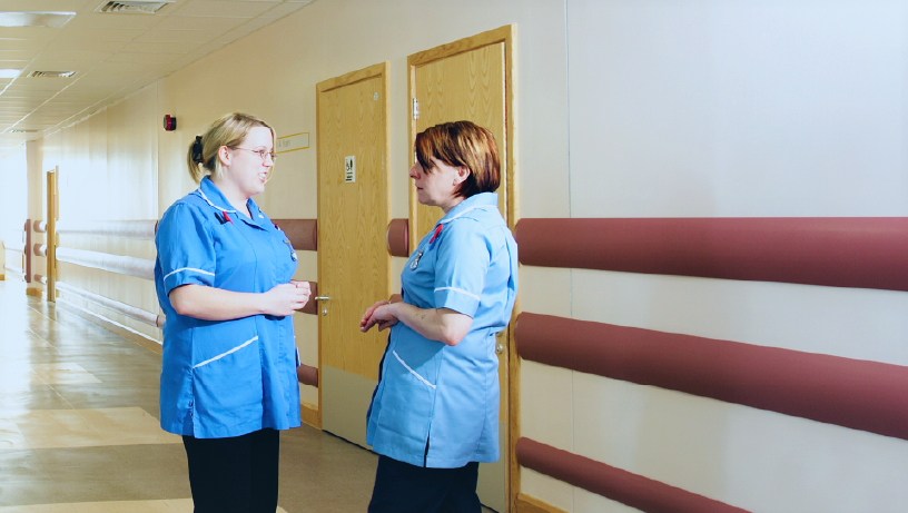 Two individuals in blue uniforms stand in a hallway of a healthcare facility with beige walls, brown handrails, and wooden doors. Their faces are obscured.