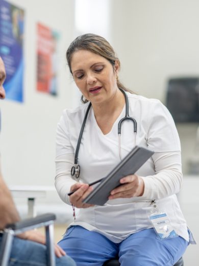 Healthcare professional in scrubs with a stethoscope, holding a tablet and speaking with an older male patient in a clinical setting.