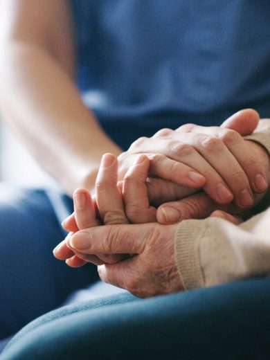 person in blue scrubs gently holding the hand of another person wearing a beige sweater, conveying support and care.