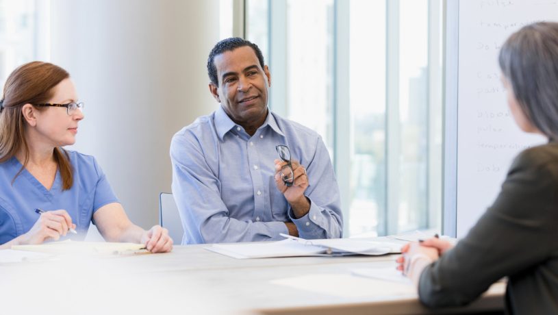 Three people in a meeting room, two in professional attire and one in scrubs, seated at a table with a whiteboard in the background.