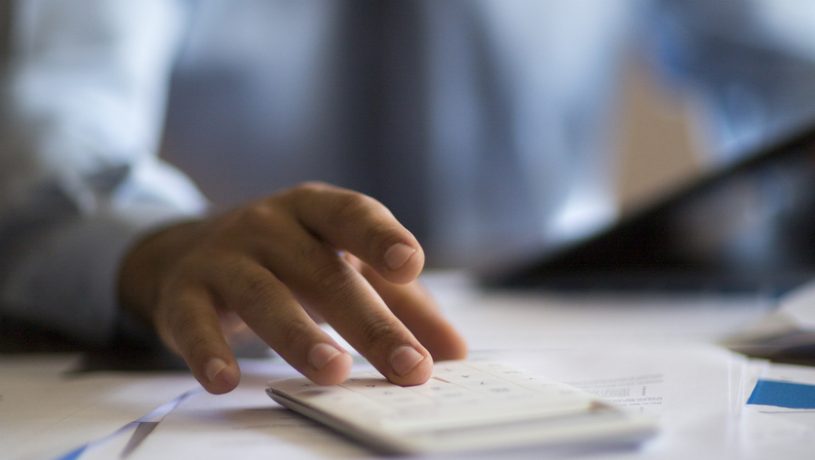 person in a blue shirt and tie using a calculator at a desk covered with papers and charts